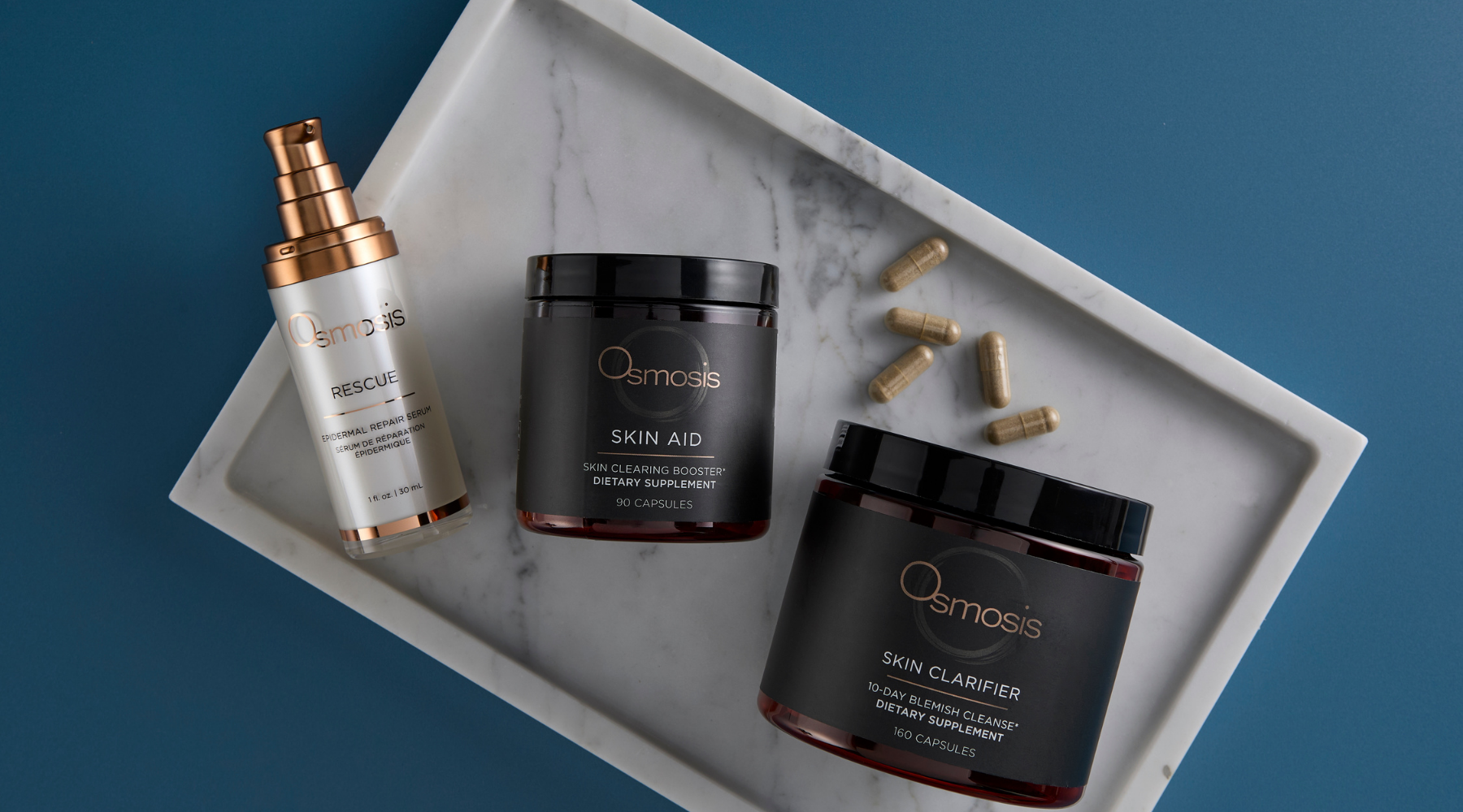 Two black twist top jars, a white skincare bottle with a pump and a supplements laying on a white marble tray on a blue background