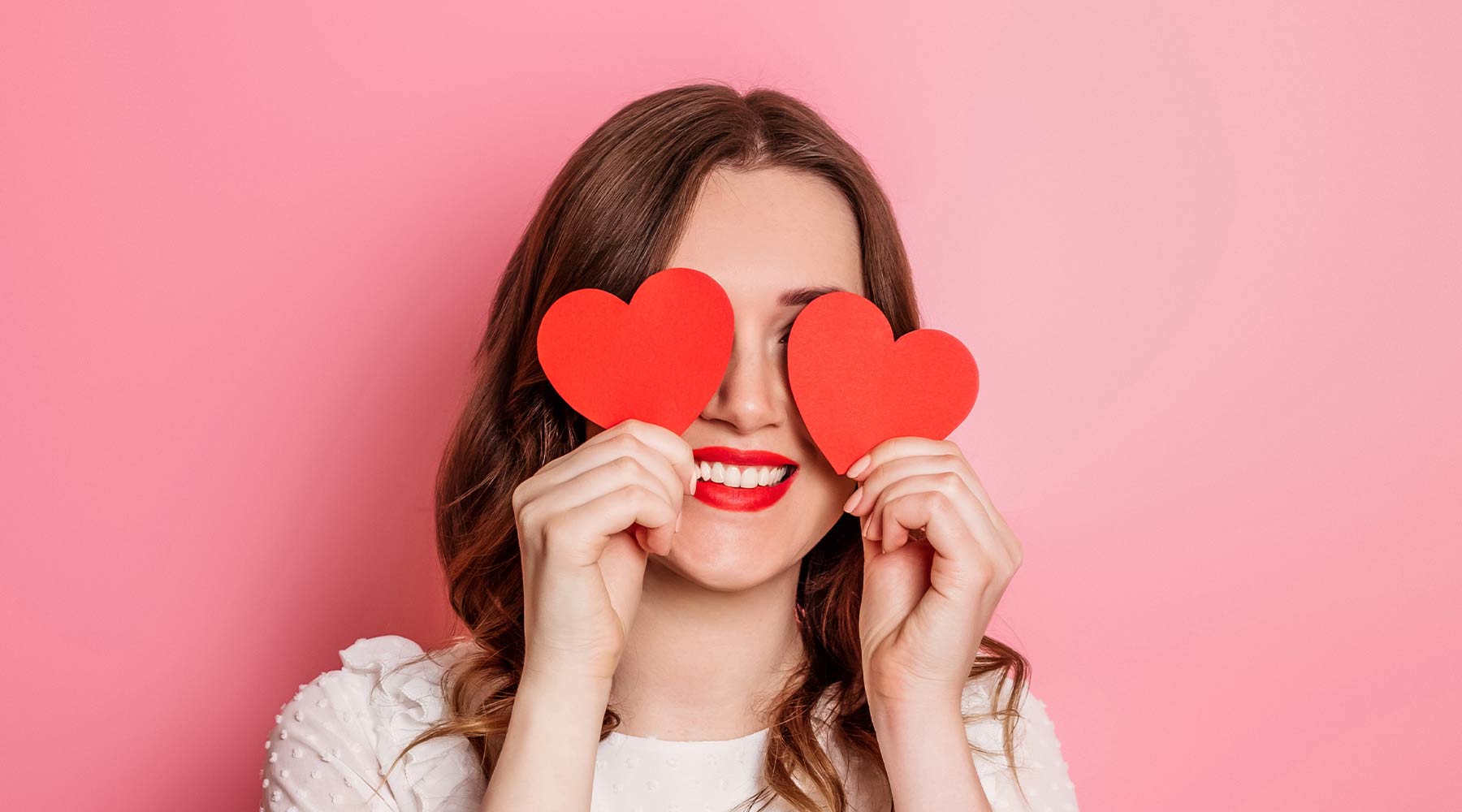 Women with brown hair in front of a pick background holding two red hearts in front of eyes