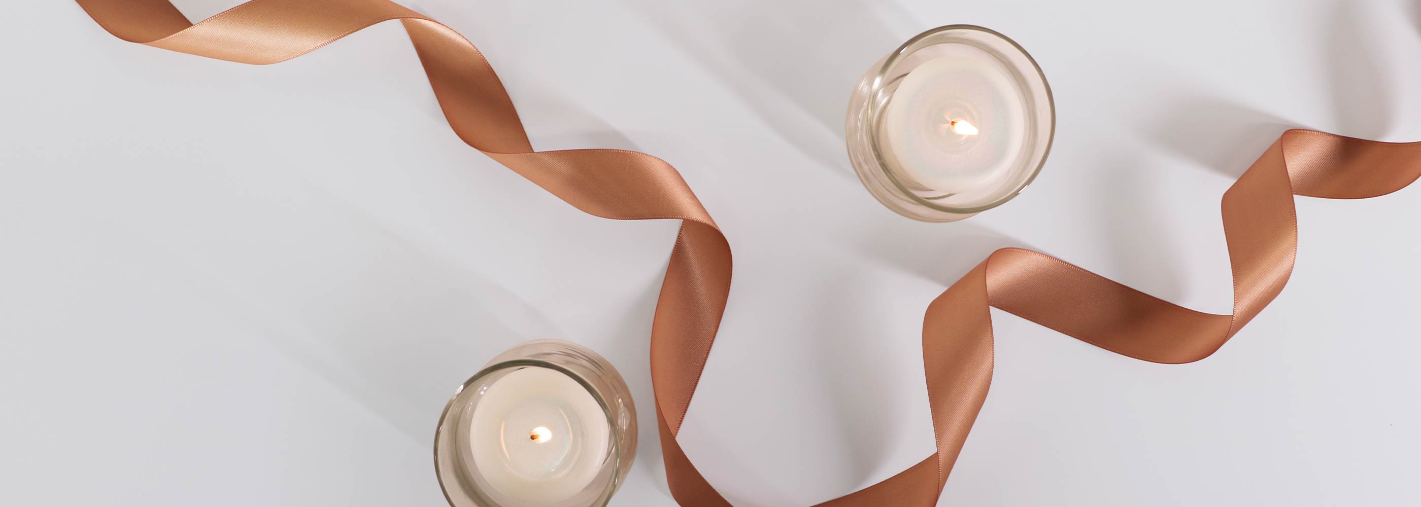 2 white candles in glass votives with a copper ribbon across the scene on a white background