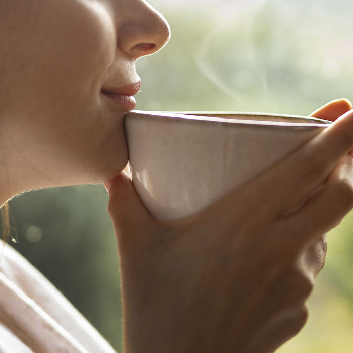 Person holding a white mug close to their face with a blurred natural background