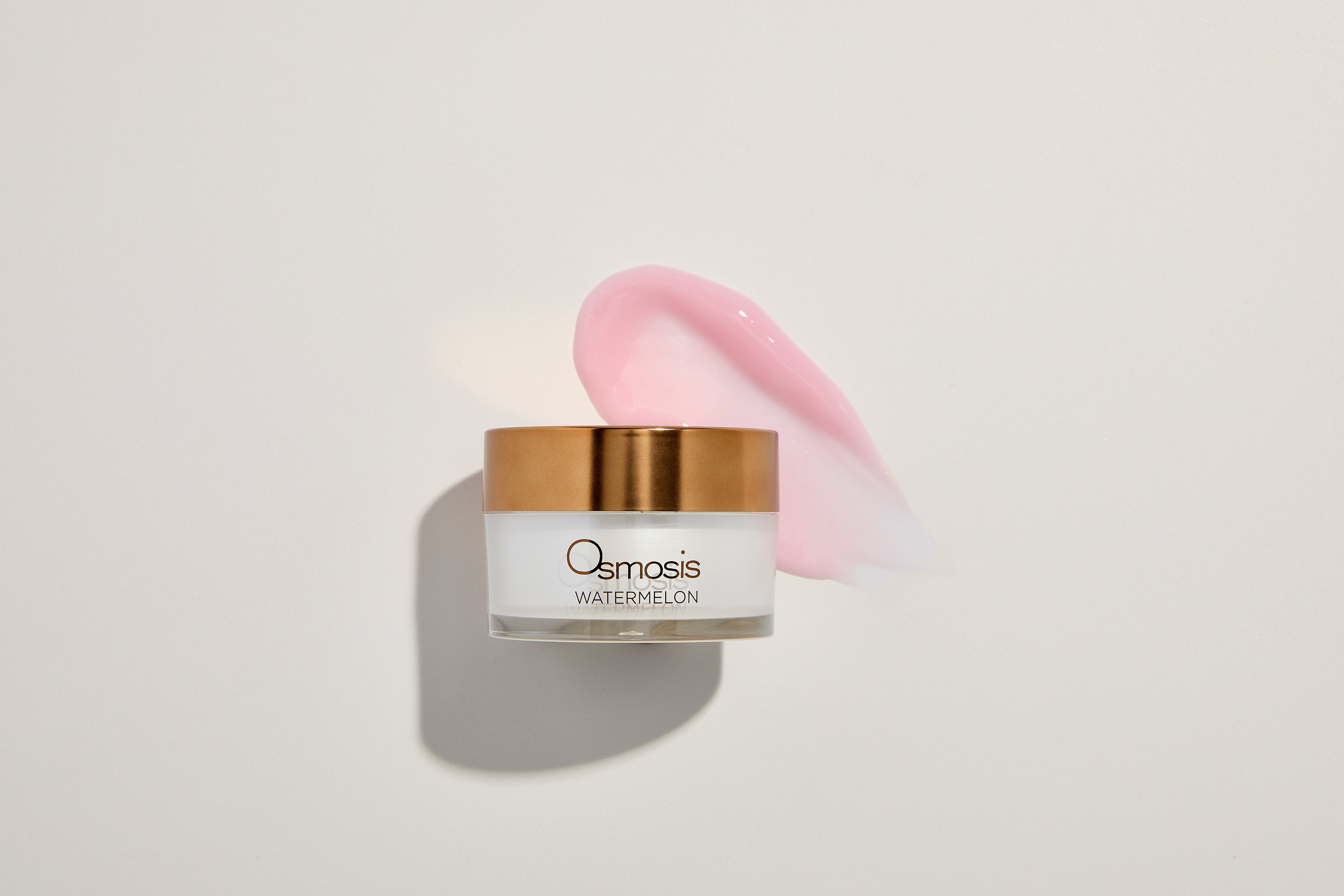 White jar with copper lid on a white background with a pink product spread behind it.